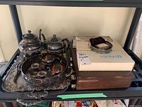 Overview of silver-plated tea and coffee pots, serving trays, cutlery boxes on a black shelf