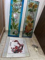 Photo showing two vertical stained glass style window panels leaning against the wall and the framed Asian needlepoint artwork laid flat along with a glass night light plug.