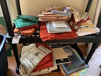 Overview of assorted table linens including pot holders, table runners, placemats, and tablecloths stacked on shelving unit.