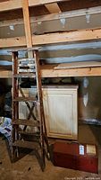 Vintage wooden ladder with paint splatters and weathered wood leaning against shelving, beside a cabinet and toolbox on the floor.