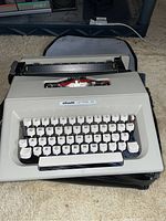 Front view of the Olivetti Lettera 30 portable typewriter showing keyboard and body condition, resting on top of its case, placed on carpeted floor.