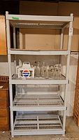 Front view of white heavy duty plastic utility shelf with four shelves. Second shelf contains glass jars and a white Pepsi carrier box.