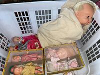 Photo showing large doll wrapped in cream blanket with multiple smaller dolls dressed in vintage clothing inside a white plastic basket and two boxed doll sets.