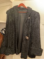 Front view of the black vintage Persian lamb fur jacket hanging on a wooden hanger with visible button and curved fur texture.