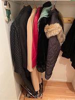 Photo showing six winter coats and jackets hanging together, mostly wool and quilted styles, varying colors including dark patterned, beige, red, black, and brown. Fur trims visible on some outerwear.