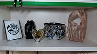Photo of shelf containing all lot items including framed Native American art tile, black soapstone figure, art glass heart, pottery bowl, and carved soapstone figures.