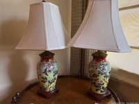 Pair of Asian ginger jar lamps with floral and bird painted ceramic bases and white fabric shades on wooden stands.