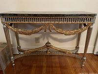Front view of console table showing black or grey top and gold ornate carved wooden legs and apron with decorative leaf garlands and finial stretcher