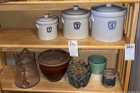 Three cream-colored numbered dry goods crocks on a wooden shelf with assorted pottery pots and a clear glass pitcher below.