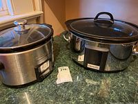Two crock pots on green countertop. Left: Oval Crock-Pot with black lid and stainless steel base. Right: Round Cuisinart slow cooker with digital control panel.