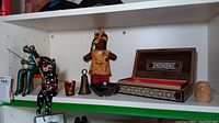 Photo showing a shelf with wooden cat and frog figurines holding fishing poles, a doll in fabric and wood dress, a brass bell, an ornamental bowl, a wooden keepsake box with detailed inlay, and a small carved wooden head.