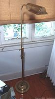 Full view of the brass floor lamp showing its height, base, and adjustable pole against a window background.