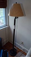 Full view of floor lamp with beige shade, tall metal shaft, and black base near window.