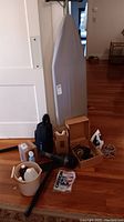 Photo of vacuum cleaner, ironing board, bucket with supplies, iron, and attachments arranged on floor