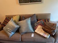 Throw pillows in various colors and patterns arranged on a beige sofa alongside folded table linens including napkins, placemats, and a tablecloth.