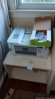 Brother MFC-J995DW printer on light wooden wheeled cart, extra ink cartridges in box on top