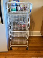 Full view of the white metal wire rack with multiple wire basket drawers, some containing household items like disinfecting wipes and canned goods.