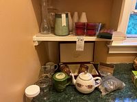 Photo of shelf showing teapot, green tea bag holder, various glass and ceramic vases, red candle holders, and glass canisters