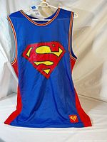 Full front view of blue sleeveless Superman basketball jersey with red side panels and multicolor striped neck and armholes on hanger.
