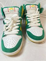 Front angled close-up view of white, green, and yellow Nike SB Dunk High sneakers showing upper details and lacing.