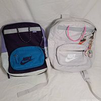 Two Nike backpacks side by side on white background, one black with blue pocket and purple accents, the other light grey with pink bag accents and keychain attached