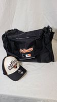 Front view of black San Francisco Giants embroidered travel duffel bag with front pocket and MLB logo, placed next to a black and white baseball cap embroidered with '2010 Champions'.