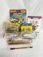 Photo showing all lot contents including crayons box, pens and markers in plastic cases, wooden brushes, banana pouch, Hello Kitty case, USA map magnets, and learning book.