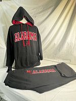 Front view of black Alabama pullover hoodie on mannequin with red inner hood and matching pants with logo placed on table