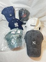 Six caps shown in a cluster on a white background including blue, navy, gray New York Yankees caps, white California cap, gray USA flag cap, and green Los Angeles cap.