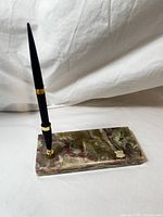 Rectangular onyx base with natural green and reddish marbling, gold-tone pen holder, black tapered pen inserted.