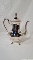 Photo of the vintage silver plated coffee pot front and side view showing polished reflective body, ornate finial, curved spout, and sculpted feet.