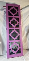 Closed view of a tall purple wooden wall hanging jewelry cabinet with a mirrored front door. The mirror has decorative geometric cut-out designs.