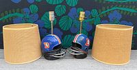 Photo showing two blue vintage Denver Broncos helmet lamps with beige woven lamp shades, cords wrapped around the helmets, lamp fixtures visible, helmets positioned on a concrete surface with a leafy patterned background.