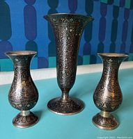 Full view of three brass vases with etched peacock and vine motifs on a turquoise surface with a blue patterned background.