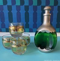 Four Culver Prado Green Roly Poly glasses with gold decorative bands and a Royal Hilland KMD pewter capped green glass decanter with stopper on a turquoise surface.