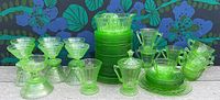 Full lot of green depression glass including plates, sherbet cups, juicer, covered sugar bowl, and bowls shown against floral fabric background.
