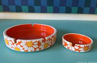 Photo showing two ceramic ashtrays, different sizes, with orange interior and white daisy floral exterior.