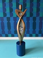 Front view of the bronze geometric sculpture showing the abstract human figure with open cutout and blue textured pattern inside, placed on a blue cylindrical base.