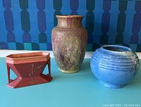 Three Roseville pottery vases on blue surface against geometric wallpaper. Left is red rectangular Carnelian II vase, middle is tall plum purple Carnelian II vase with drip glaze and chips, right is round blue Tourmaline vase with ridges.