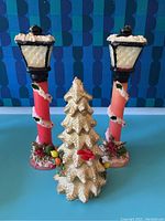 Front view of two holiday street lamp candles and one Christmas tree candle on blue surface with patterned blue backdrop.