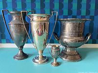 Four tarnished silver trophies of different sizes including a small two-handled 1927 Pretty Baby Show trophy with engraving, a large 1984 District Man of the Year trophy bowl, and two other large urn/cup-shaped trophies, all on blue surface with blue-patterned wall background.