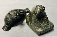 Two soapstone carvings, a dark seal with carved flippers and a greenish bear, shown from front and side angles on grey fabric background.
