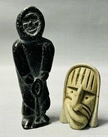 Front view of a dark soapstone standing Inuit figure and a lighter stone abstract face carving, showing detailed carving and finish.