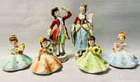 Full view of all six porcelain figurines on neutral background. Two large standing adult figures in back, four smaller seated female figures in front holding flowers.