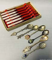Full view of 6 dessert knives with mother-of-pearl handles in box along with 6 assorted silver plate collectible spoons.