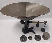 Front and top view of antique cast iron balance scale with large oval metal weighing pan and four metal weights placed in front.