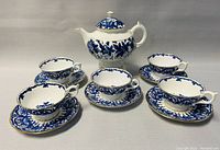 Full view of the covered teapot placed centrally with five matching teacups and saucers arranged around it, showcasing the blue floral design and gold edging on white bone china.