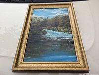 Framed 19th century landscape painting showing a river and trees, with gold textured frame, photographed lying flat.