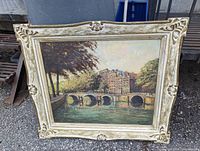 Full front image of the framed oil painting depicting a river with stone bridge and buildings.