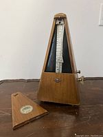 Front view of wooden mechanical metronome with removable cover placed beside it, showing tempo scale and pendulum arm.